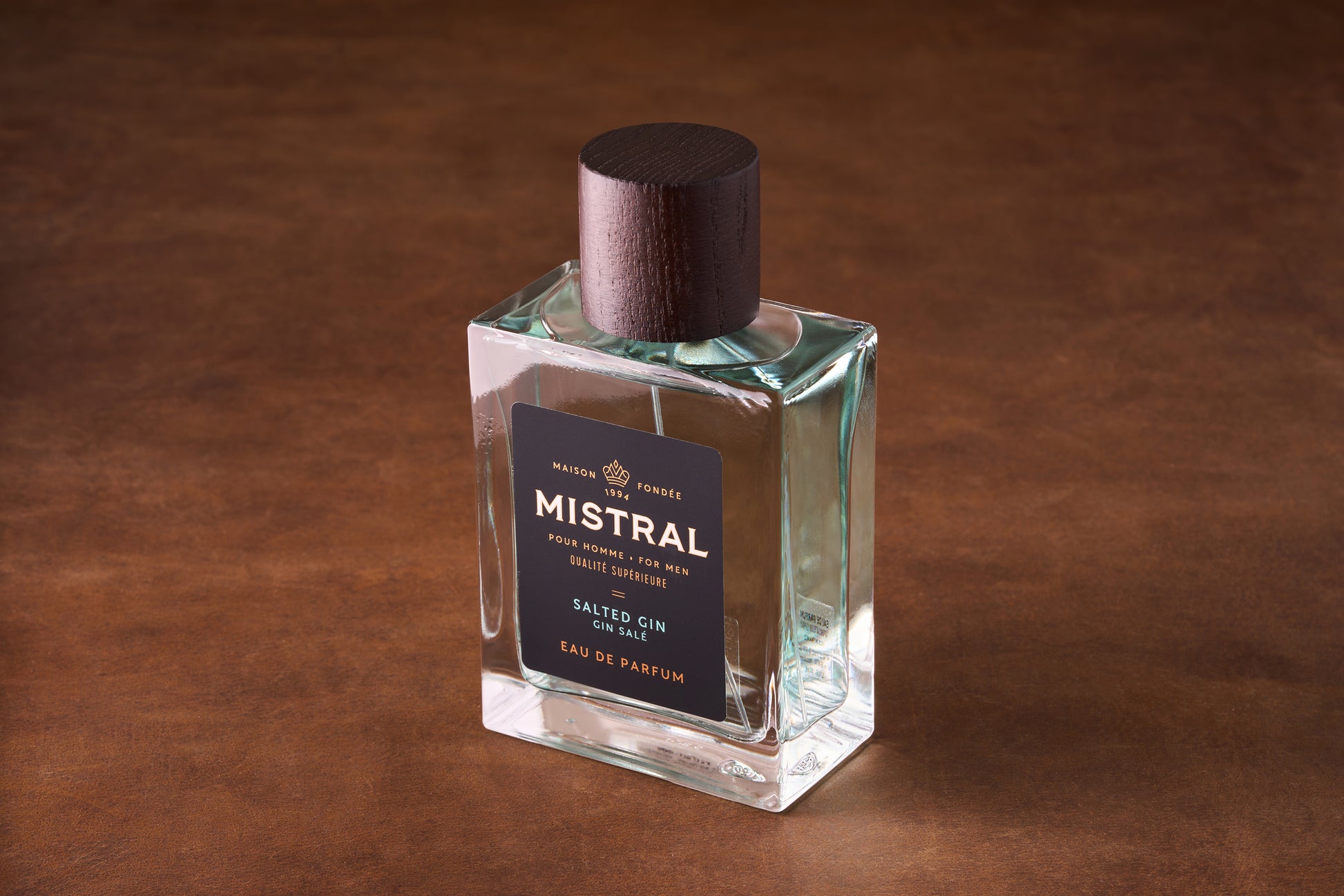 A rectangular glass bottle of Mistral Salted Gin Eau de Parfum 100ml with a dark wooden cap rests on a brown surface; its black label displays elegant white and gold text inspired by Provence perfume.
