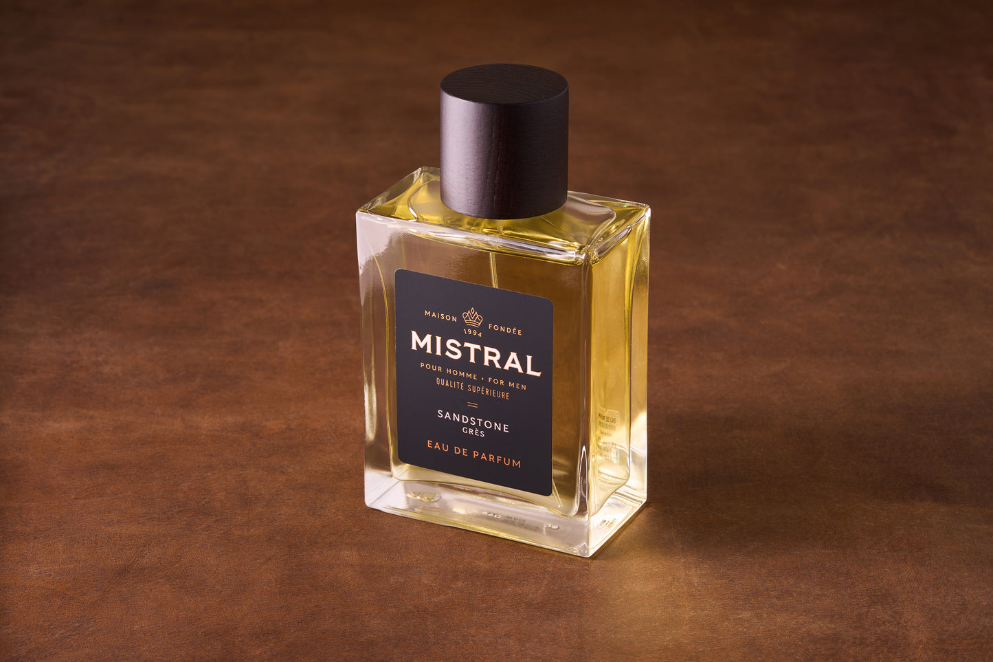 A rectangular glass bottle of Mistral Sandstone Eau de Parfum 100ml sits on a brown surface, showcasing pale yellow liquid. It features a black label with gold text and a dark cap.