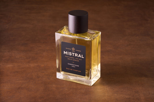 A rectangular glass bottle of Mistral Sandstone Eau de Parfum 100ml sits on a brown surface, showcasing pale yellow liquid. It features a black label with gold text and a dark cap.