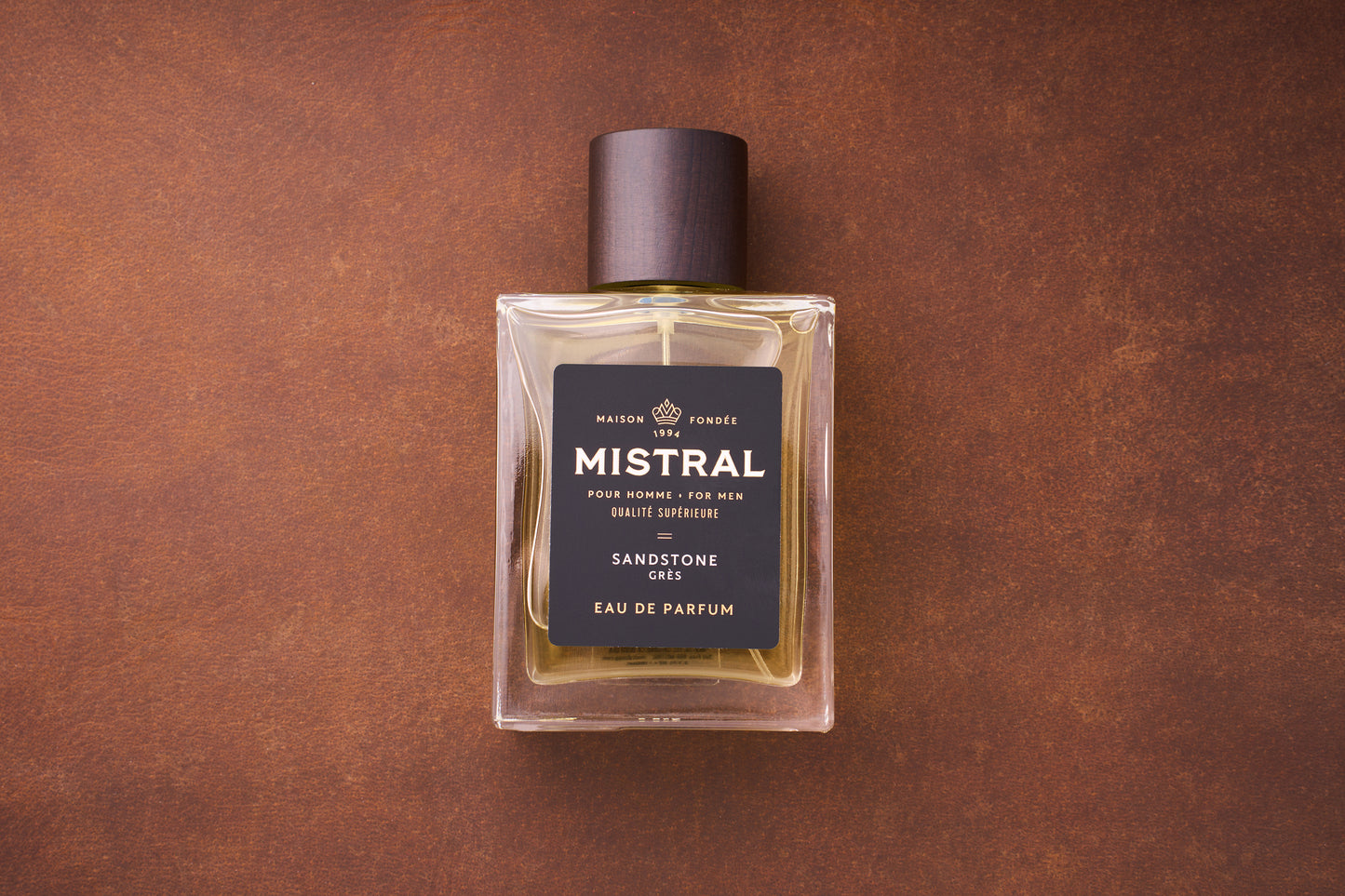 A clear glass bottle of Mistral Sandstone Eau de Parfum 100ml, a luxury French men’s fragrance with a dark brown cap and black label, sits on a brown textured surface, reflecting the essence of Provence.