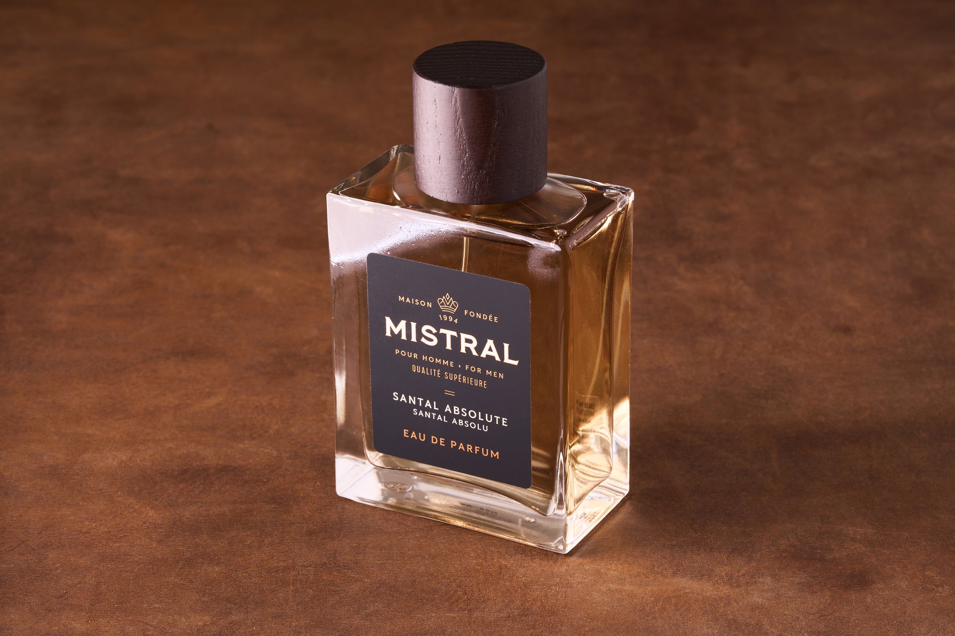 A clear glass bottle of Mistral Santal Absolute Eau de Parfum 100ml with a dark wooden cap rests on a brown surface. The label reads "Mistral Pour Homme" and showcases its exquisite French Nepalese sandalwood fragrance.