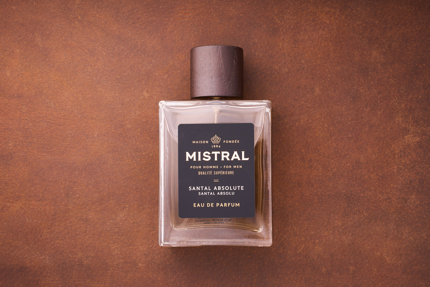 A rectangular glass bottle of Mistral Santal Absolute Eau de Parfum 100ml, a French men’s fragrance with a dark wooden cap, rests on a brown textured surface. The label features black and gold accents, highlighting Nepalese sandalwood.