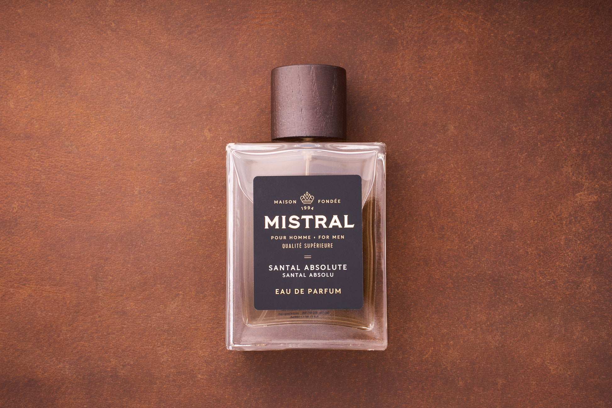 A rectangular glass bottle of Mistral Santal Absolute Eau de Parfum 100ml, a French men’s fragrance with a dark wooden cap, rests on a brown textured surface. The label features black and gold accents, highlighting Nepalese sandalwood.