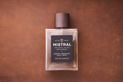 A rectangular glass bottle of Mistral Santal Absolute Eau de Parfum 100ml, a French men’s fragrance with a dark wooden cap, rests on a brown textured surface. The label features black and gold accents, highlighting Nepalese sandalwood.