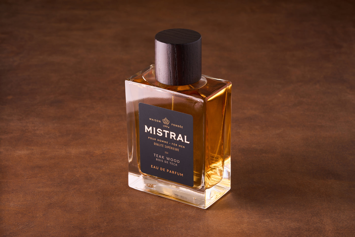 The Mistral Teak Wood Eau de Parfum 100ml, in a rectangular glass bottle with amber liquid, a dark wood cap, and black and gold label, channels midcentury style and fine French fragrance on a brown surface.