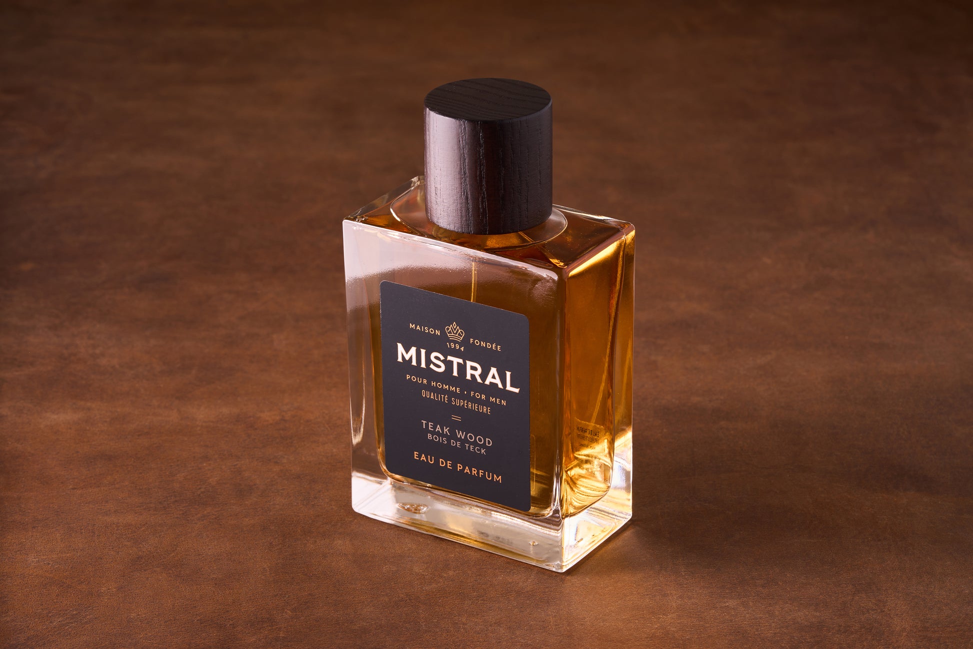 The Mistral Teak Wood Eau de Parfum 100ml, in a rectangular glass bottle with amber liquid, a dark wood cap, and black and gold label, channels midcentury style and fine French fragrance on a brown surface.