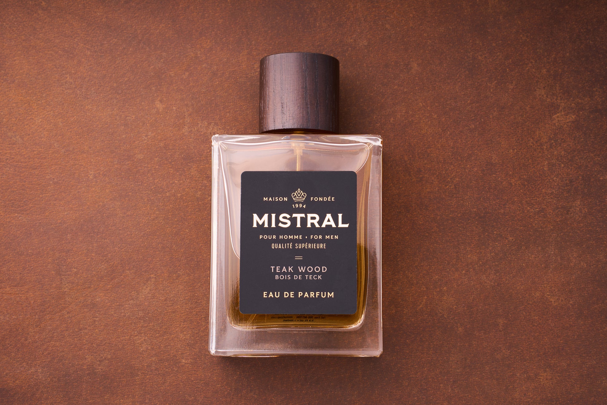 A 100ml bottle of Mistral Teak Wood Eau de Parfum for men, topped with a wooden cap, sits on a brown textured surface. The French label features the brand and scent details in gold and white text.