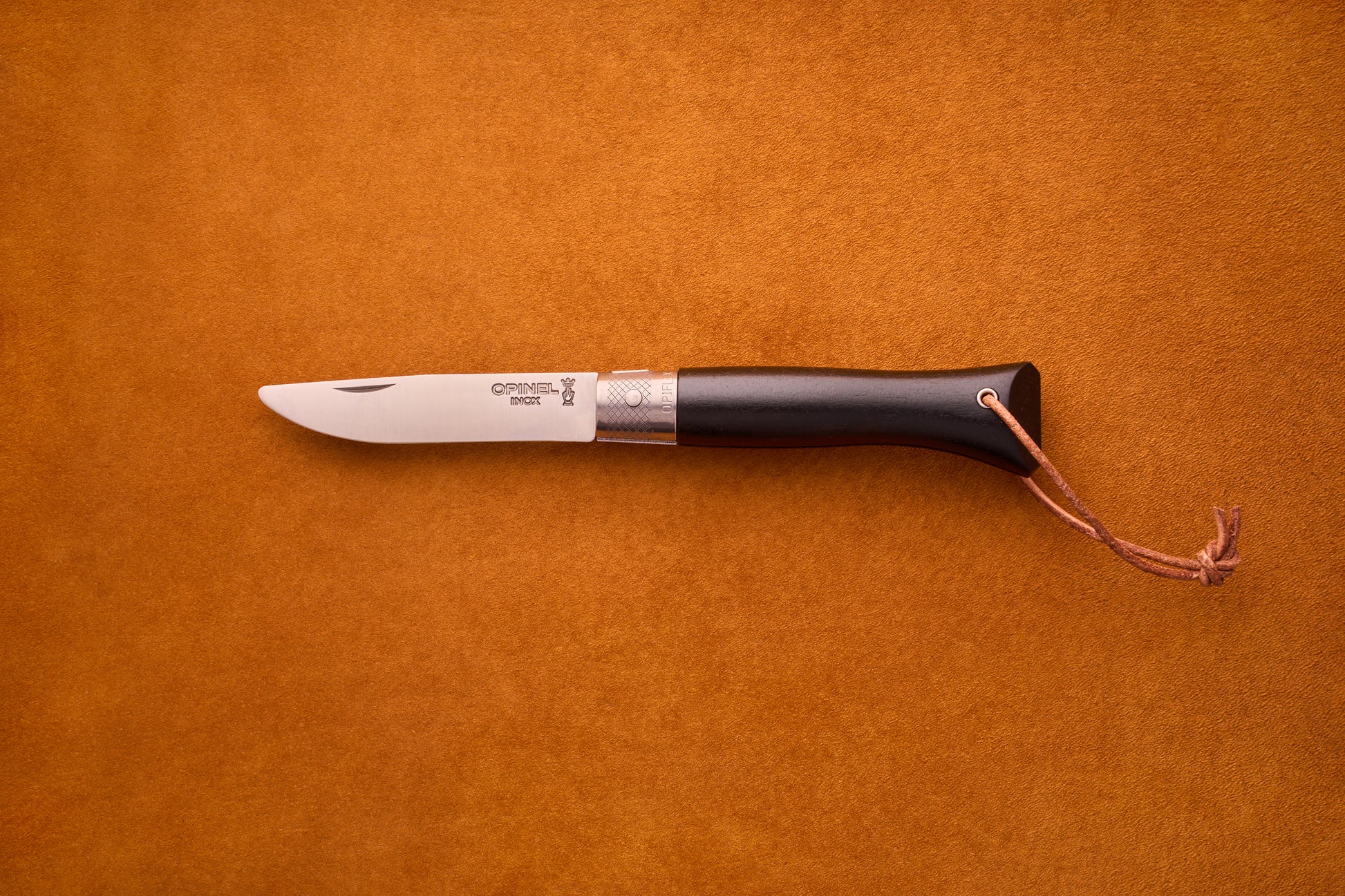 The Opinel Neo6, a folding pocket knife featuring an ebony wood handle, silver bolster, and a small leather lanyard, is displayed on a brown suede background—an elegant choice for everyday carry.