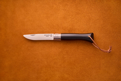 The Opinel Neo6, a folding pocket knife featuring an ebony wood handle, silver bolster, and a small leather lanyard, is displayed on a brown suede background—an elegant choice for everyday carry.