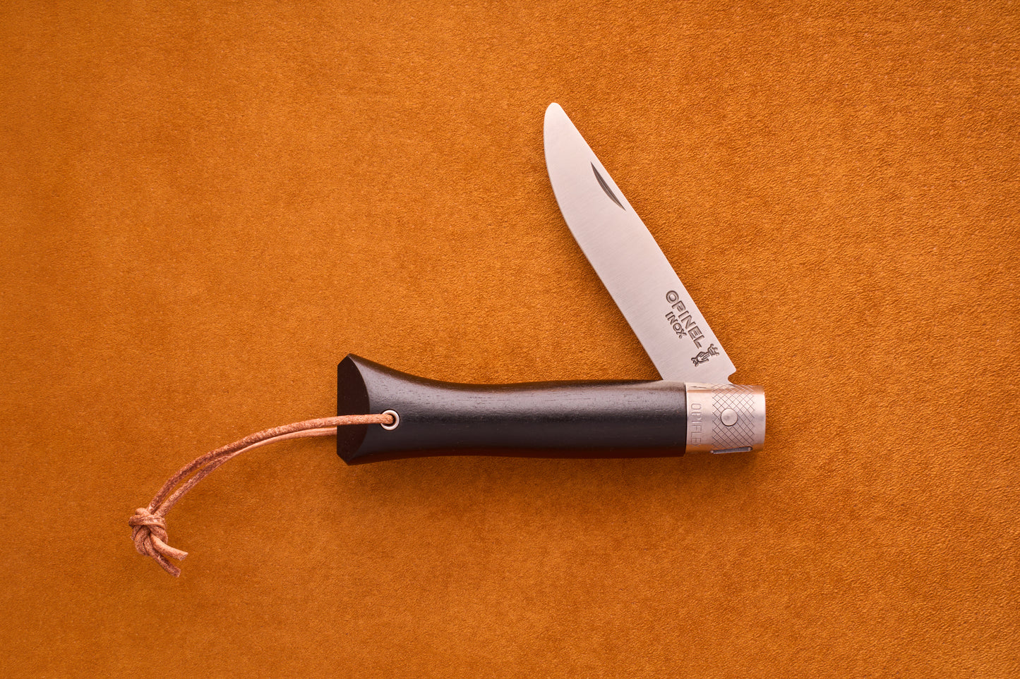 An Opinel Neo6 pocket knife with an elegant ebony wood handle and a leather lanyard is open and placed on a textured orange surface, making it both a stylish and practical choice for everyday carry.