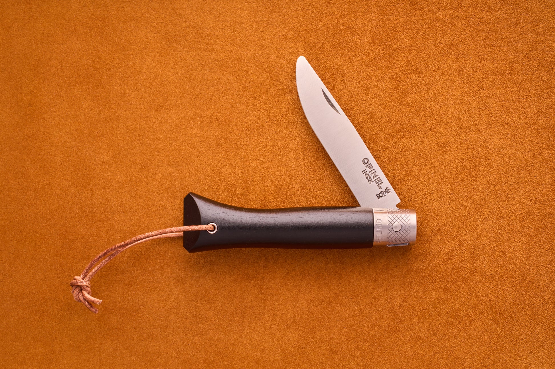 An Opinel Neo6 pocket knife with an elegant ebony wood handle and a leather lanyard is open and placed on a textured orange surface, making it both a stylish and practical choice for everyday carry.