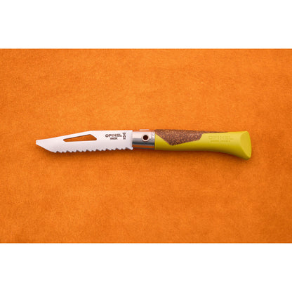 An Opinel Neo 7 Alpine kitchen knife with a green and wood handle is placed on an orange textured surface. The serrated blade, inspired by the classic Opinel pocket knife design, features a slot near the tip and displays the Opinel branding.