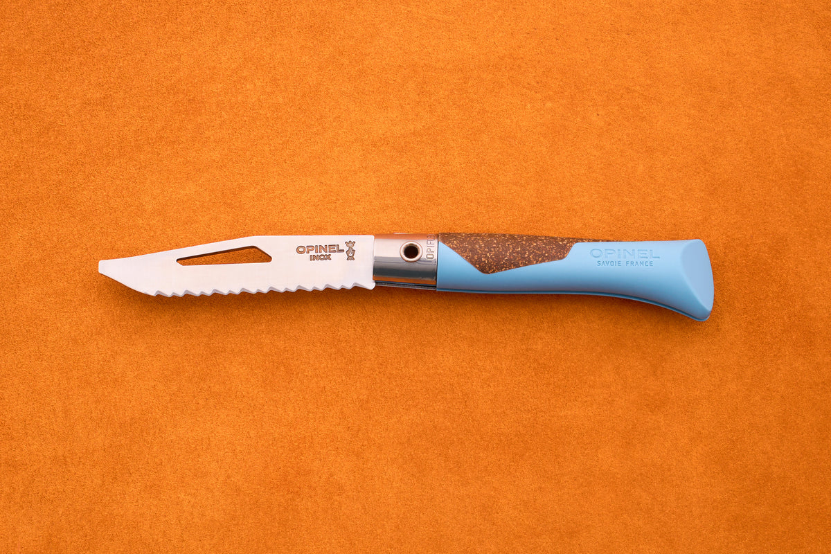An Opinel Neo 7 Alpine kitchen knife with a blue handle rests on an orange surface. The partially open blade, marked with the Opinel branding and featuring a slot near the tip, displays the signature serrated edge inspired by reliable mountain tool designs.