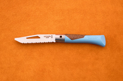An Opinel Neo 7 Alpine kitchen knife with a blue handle rests on an orange surface. The partially open blade, marked with the Opinel branding and featuring a slot near the tip, displays the signature serrated edge inspired by reliable mountain tool designs.