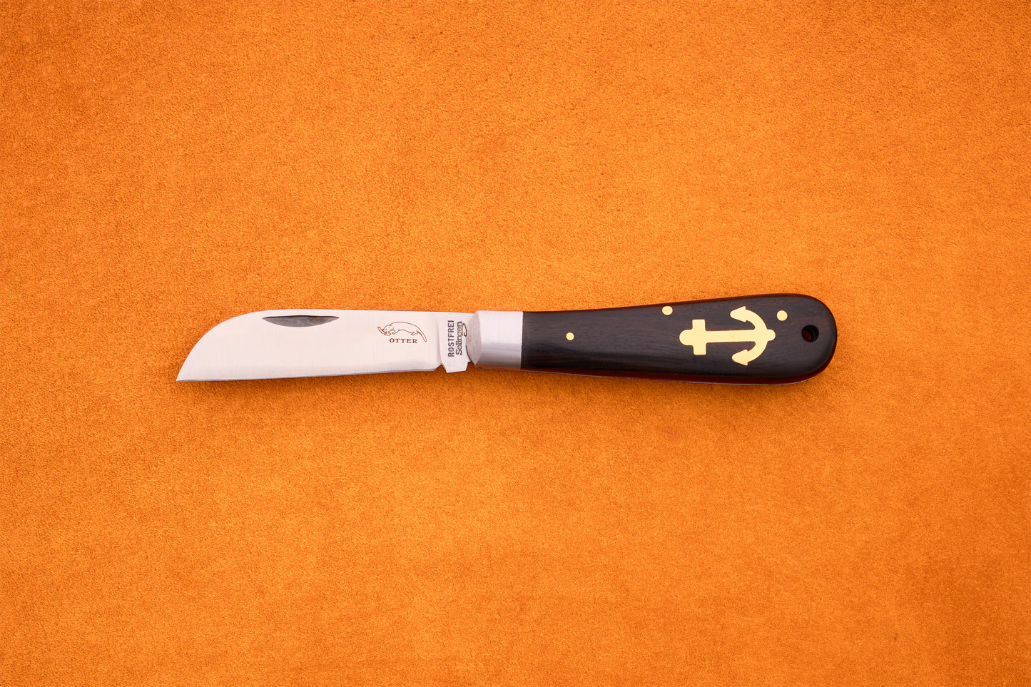 The Otter-Messer Anchor Grenadilla, a folding knife with a curved blade and a dark grenadilla wood handle adorned with a gold anchor symbol, rests elegantly on an orange surface.