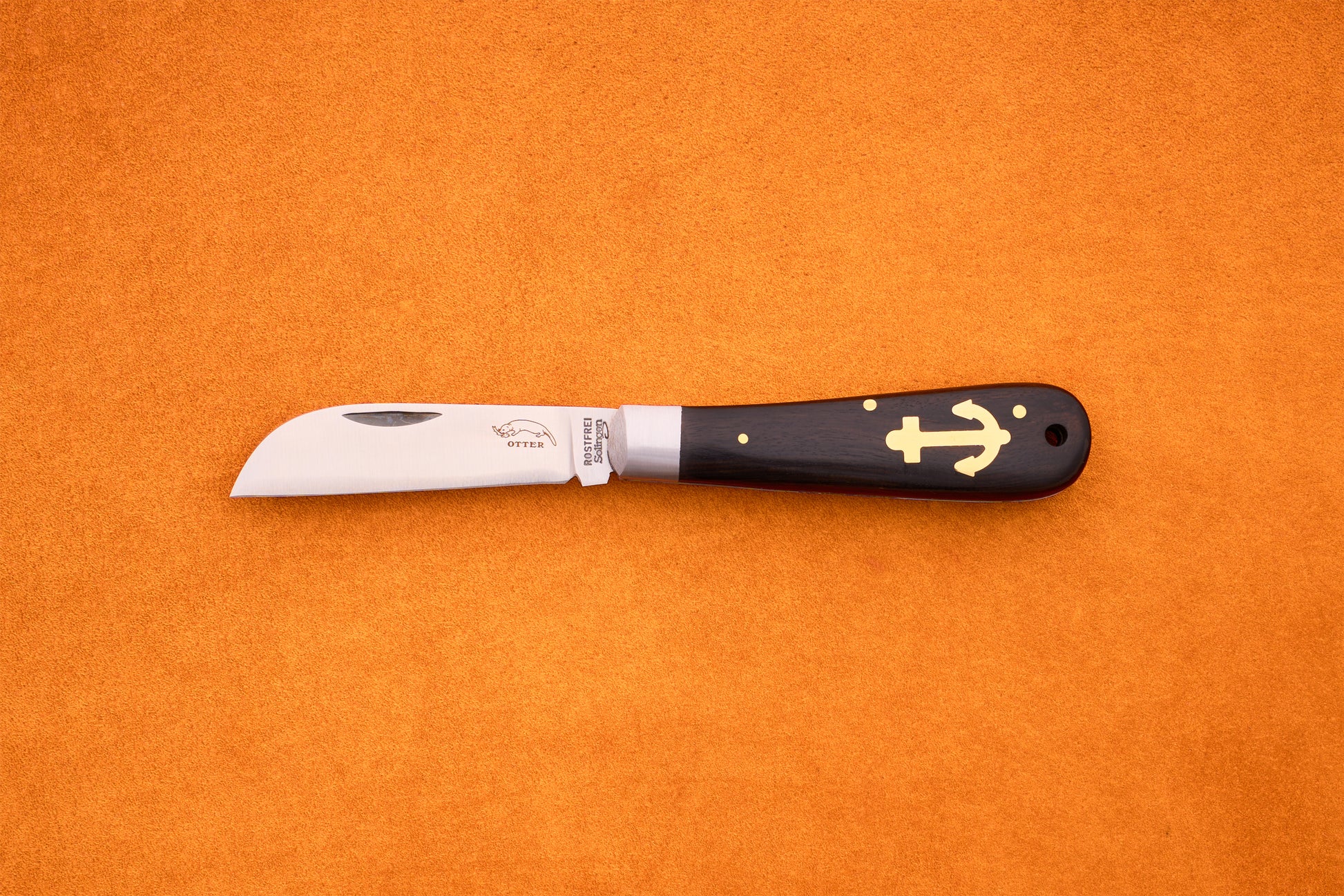 The Otter-Messer Anchor Grenadilla, a folding knife with a curved blade and a dark grenadilla wood handle adorned with a gold anchor symbol, rests elegantly on an orange surface.