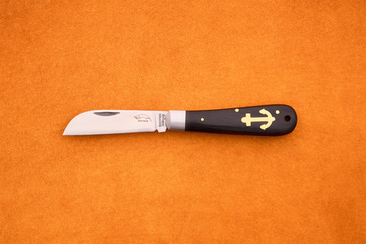 The Otter-Messer Anchor Grenadilla, a folding knife with a curved blade and a dark grenadilla wood handle adorned with a gold anchor symbol, rests elegantly on an orange surface.