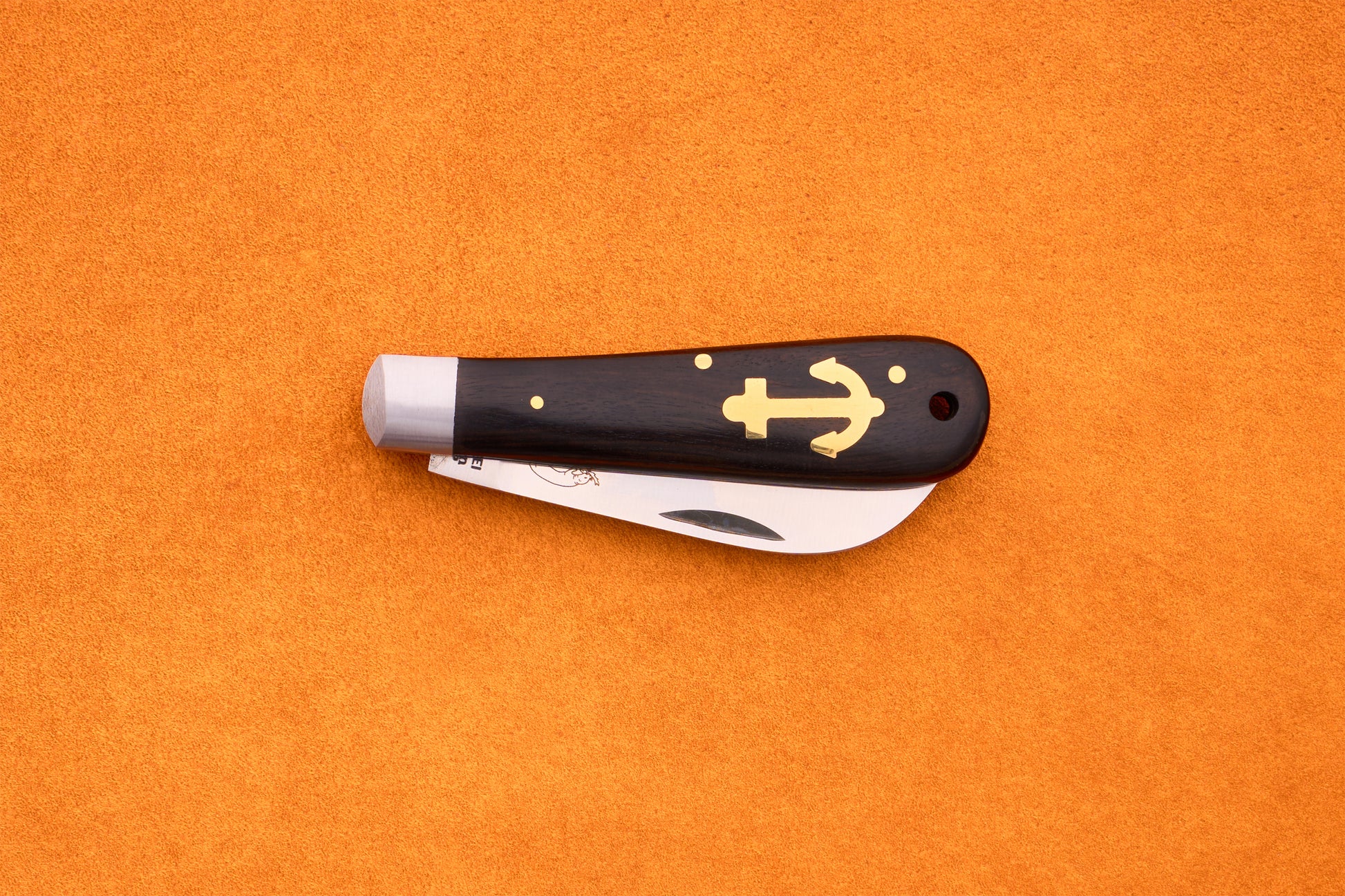 A partially open Otter-Messer Anchor Grenadilla folding knife, featuring a dark grenadilla wood handle with a gold anchor design, rests on an orange textured surface.