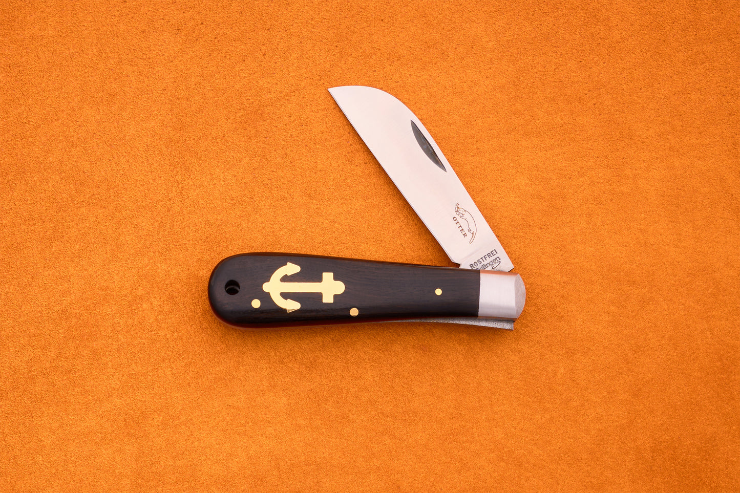 The Otter-Messer Anchor Grenadilla, a handcrafted German knife with a wooden handle and gold anchor inlay, rests open on an orange textured surface.