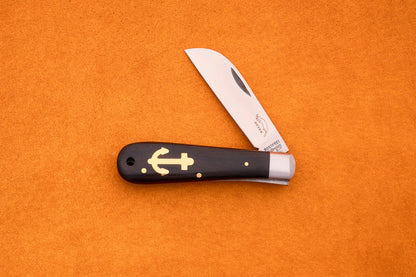 The Otter-Messer Anchor Grenadilla, a handcrafted German knife with a wooden handle and gold anchor inlay, rests open on an orange textured surface.