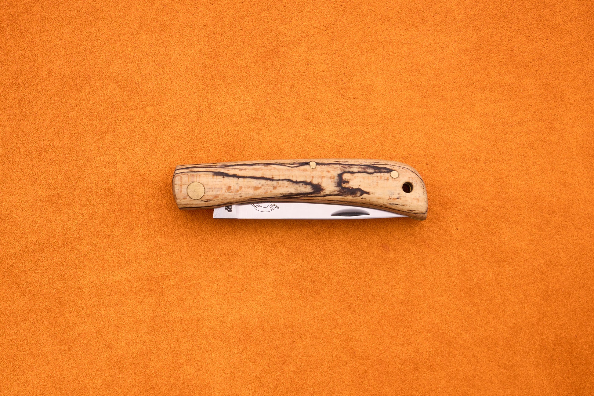 An Otter-Messer Hippe-Kniep folding pocket knife, featuring a wood-patterned handle and a partially visible blade reminiscent of a traditional farmer’s knife, is placed on an orange textured surface.