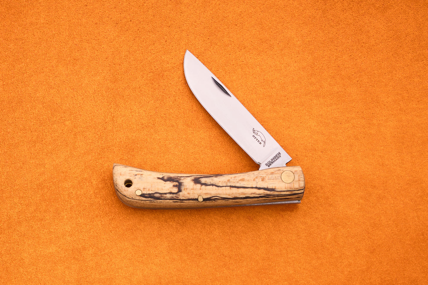 An Otter-Messer Hippe-Kniep, a handmade German knife featuring a wooden handle and a partially open silver blade, rests on an orange textured surface, evoking its heritage as a classic traditional farmer’s knife.