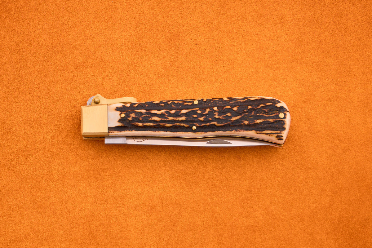 The Otter-Messer Hunting Pocket Knife, shown closed with a textured dark brown stag horn handle and brass bolster, is displayed on a bright orange background.