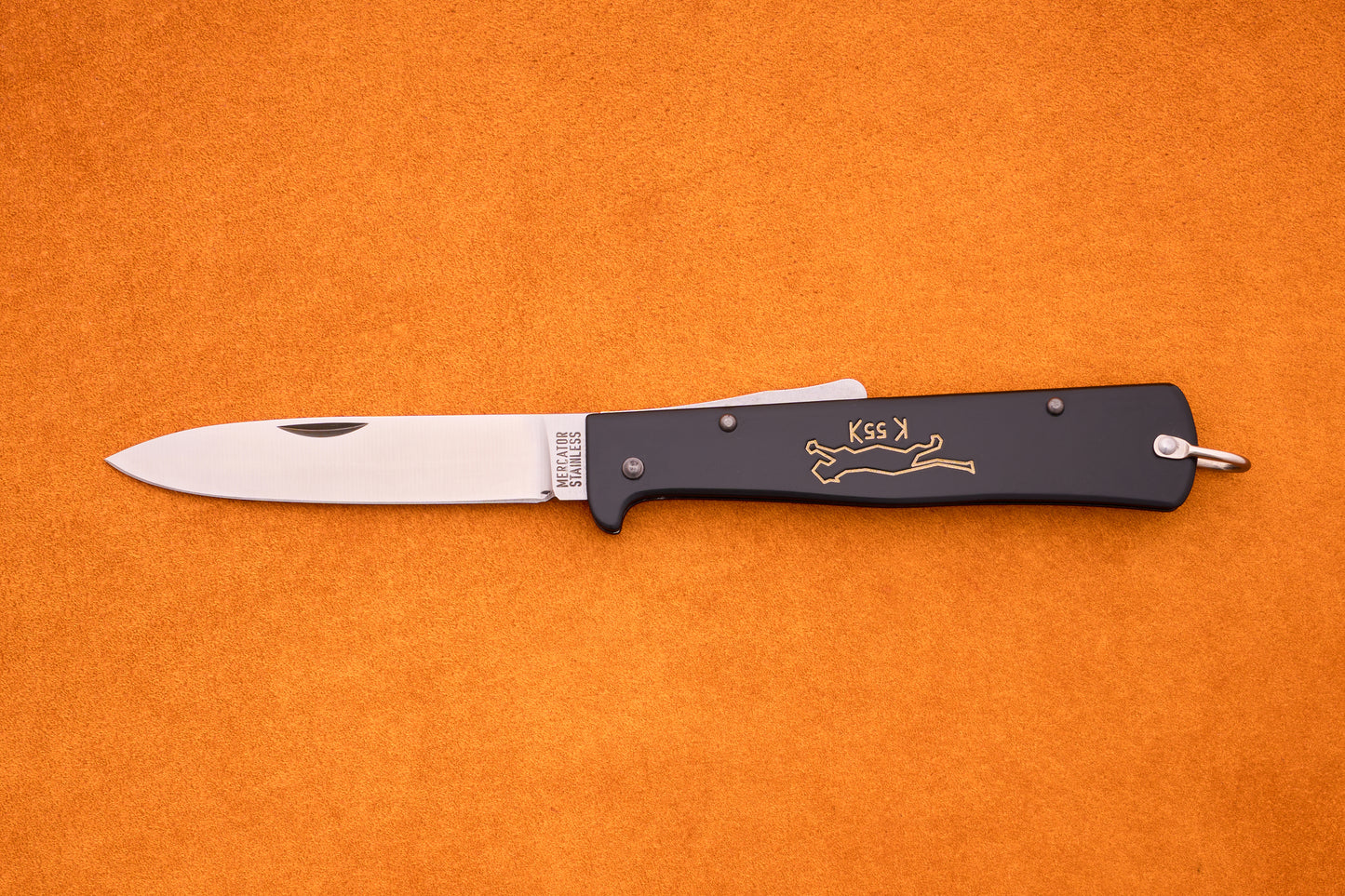 An Otter-Messer Mercator “Black Cat Knife” K55K, featuring a silver blade and black handle adorned with a gold cat design reminiscent of the iconic Mercator style, lies open on an orange surface.