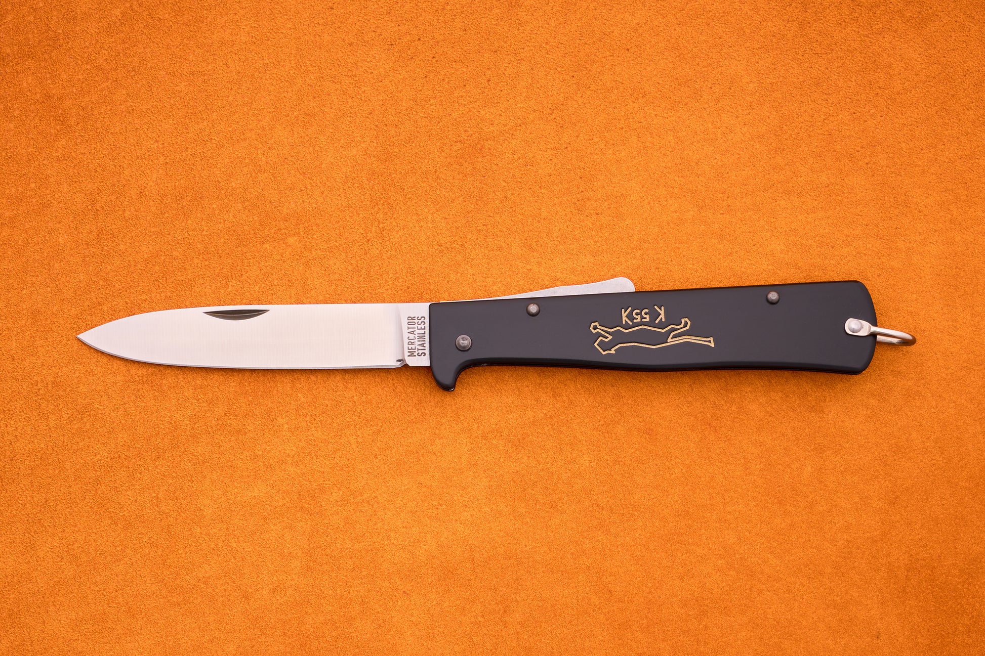 An Otter-Messer Mercator “Black Cat Knife” K55K, featuring a silver blade and black handle adorned with a gold cat design reminiscent of the iconic Mercator style, lies open on an orange surface.