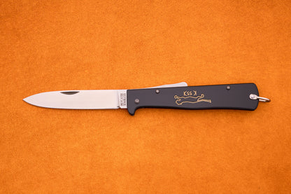 An Otter-Messer Mercator “Black Cat Knife” K55K, featuring a silver blade and black handle adorned with a gold cat design reminiscent of the iconic Mercator style, lies open on an orange surface.