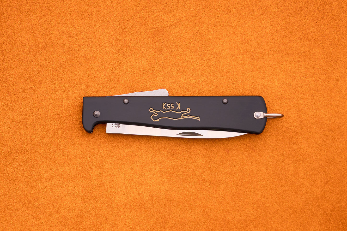 A black Otter-Messer Mercator “Black Cat Knife” K55K folding pocket knife with a partially visible silver blade, adorned with a gold running cat logo and the K55K inscription, rests on an orange textured surface. This iconic knife blends classic design with dependable performance.