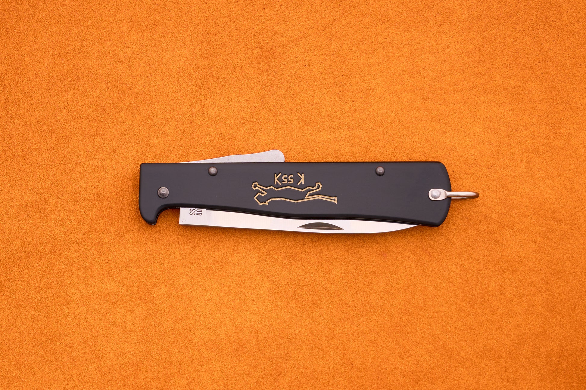 A black Otter-Messer Mercator “Black Cat Knife” K55K folding pocket knife with a partially visible silver blade, adorned with a gold running cat logo and the K55K inscription, rests on an orange textured surface. This iconic knife blends classic design with dependable performance.