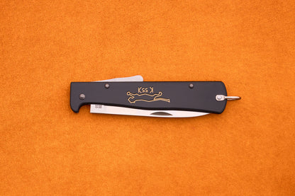 A black Otter-Messer Mercator “Black Cat Knife” K55K folding pocket knife with a partially visible silver blade, adorned with a gold running cat logo and the K55K inscription, rests on an orange textured surface. This iconic knife blends classic design with dependable performance.