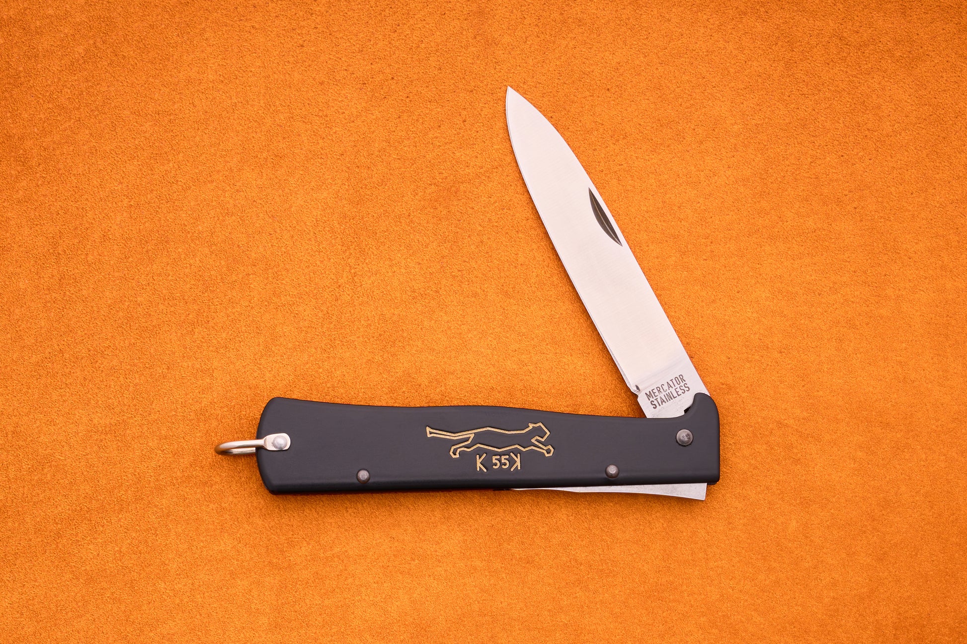 A folding pocket knife with a black handle, gold animal engraving, and a partially opened silver blade lies on an orange surface—this stylish Otter-Messer Mercator “Black cat knife” K55K showcases classic design and dependable craftsmanship.