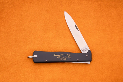 A folding pocket knife with a black handle, gold animal engraving, and a partially opened silver blade lies on an orange surface—this stylish Otter-Messer Mercator “Black cat knife” K55K showcases classic design and dependable craftsmanship.