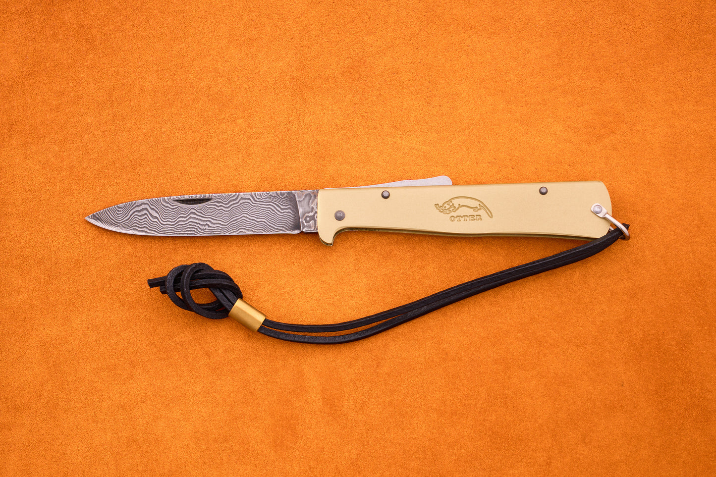The Otter-Messer Mercator Brass Damascus, a folding pocket knife crafted in Solingen, Germany, features a patterned Damascus blade, a tan handle, and a black leather lanyard, shown on an orange surface.