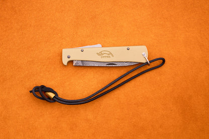 Displayed on an orange surface, the Otter-Messer Mercator Brass Damascus folding pocket knife features a beige handle, an intricately patterned blade, and a black lanyard—expertly crafted in Solingen, Germany, this knife exemplifies classic style and renowned German craftsmanship.