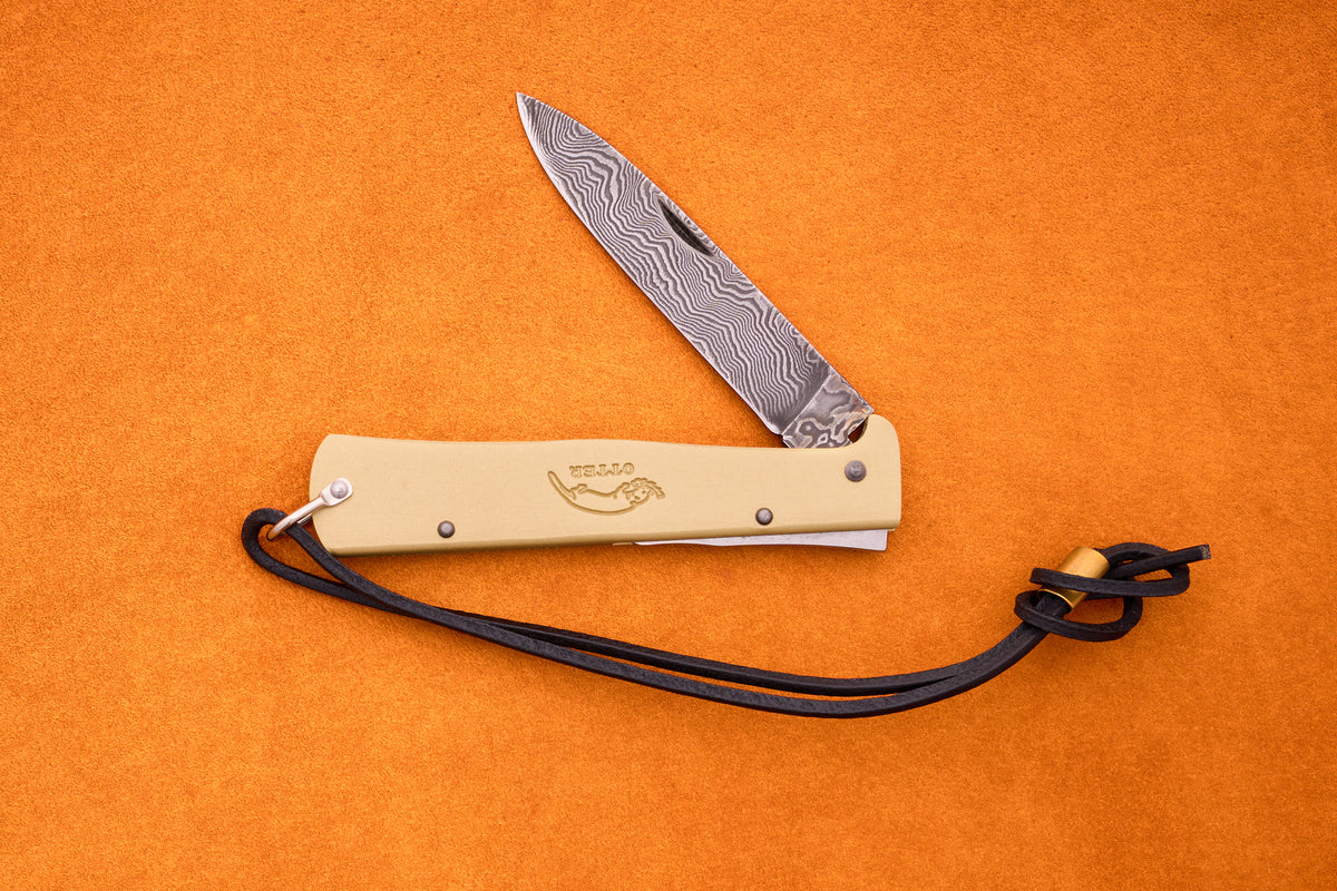 The Otter-Messer Mercator Brass Damascus folding pocket knife, expertly crafted in Solingen, Germany, features a patterned Damascus blade, a tan handle, and a black leather lanyard. Displayed on an orange surface, it exemplifies traditional design and superior craftsmanship.