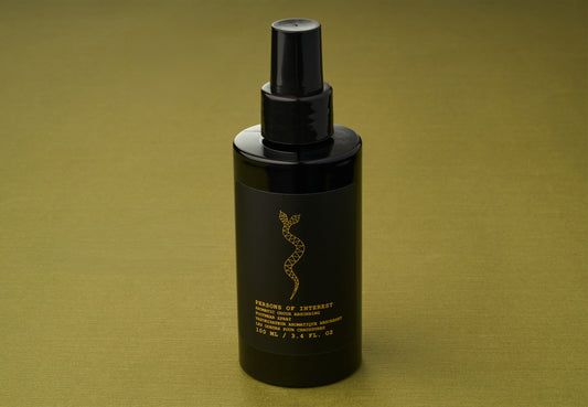 A black spray bottle labeled “Persons of Interest Aromatic Odour Absorbing Footwear Spray,” featuring gold text and a snake motif, stands on an olive green surface, hinting at an alluring fragrance inspired by rare essential oils.