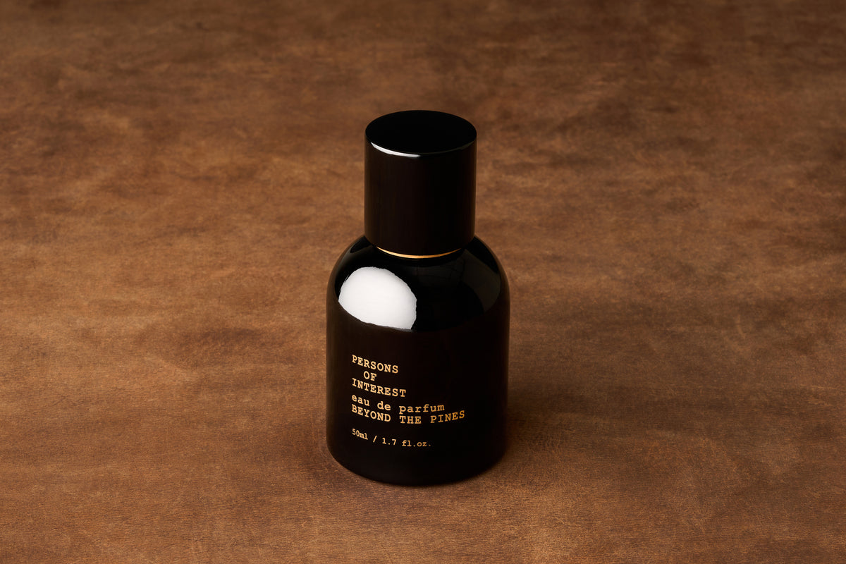 A black bottle of Persons of Interest Beyond the Pines Eau de Parfum, a unisex fragrance handcrafted in Canada, stands on a brown textured surface.