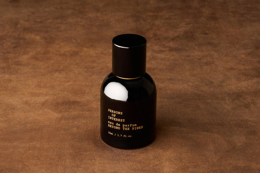 A black bottle of Persons of Interest Beyond the Pines Eau de Parfum, a unisex fragrance handcrafted in Canada, stands on a brown textured surface.
