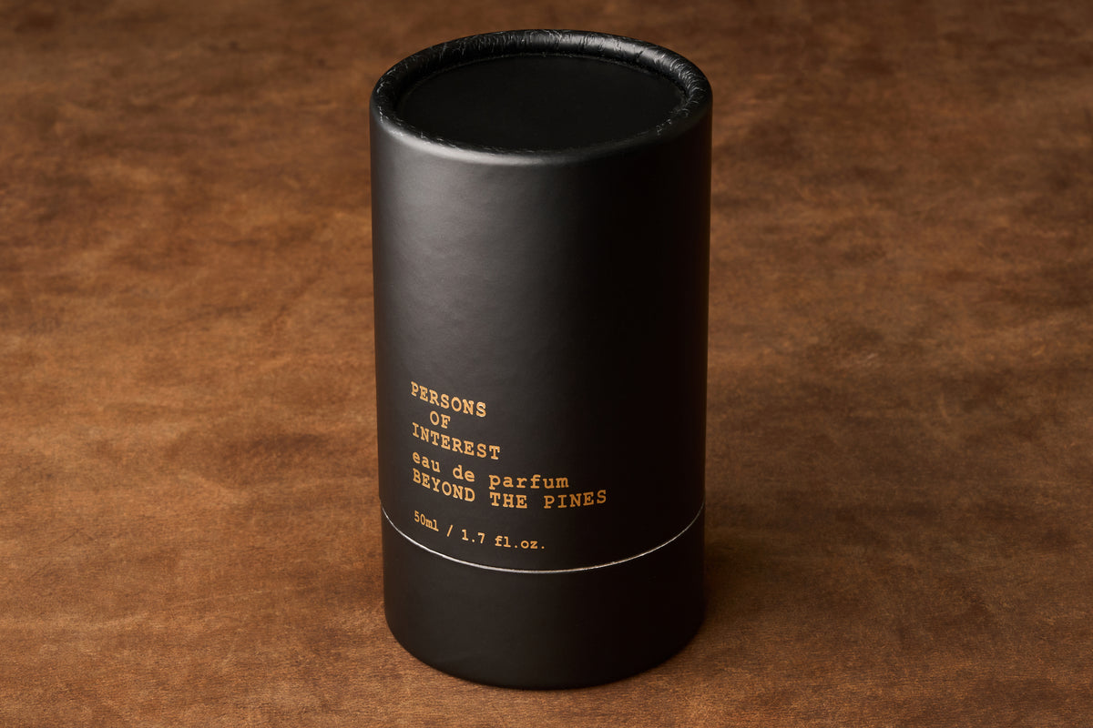 A black cylindrical perfume box labeled “Persons of Interest Beyond the Pines Eau de Parfum” in gold lettering rests on a brown textured surface, showcasing this handcrafted unisex fragrance made in Canada.