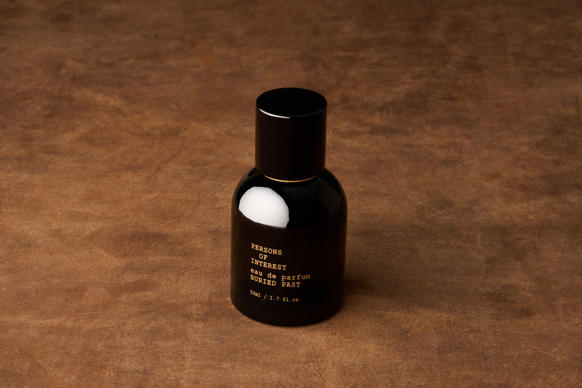A black perfume bottle labeled “Persons of Interest Buried Past Eau de Parfum 50ML” rests on a brown textured surface, illuminated by soft lighting with minimal shadows—a sophisticated presentation for this captivating unisex fragrance.