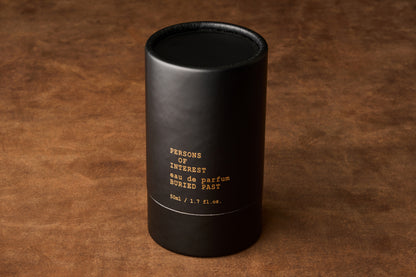 A black cylindrical container labeled Persons of Interest Buried Past Eau de Parfum 50ml / 1.7 fl.oz., a sophisticated unisex fragrance, rests on a textured brown surface.