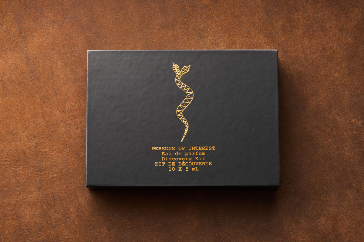 A black box adorned with a gold geometric snake design and gold lettering displaying “Persons of Interest Eau de Parfum Discovery Kit, 10 x 5ml Bottles” rests on a brown textured surface, inviting you to discover a curated collection of unisex fragrances from Persons of Interest.