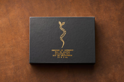 A black box adorned with a gold geometric snake design and gold lettering displaying “Persons of Interest Eau de Parfum Discovery Kit, 10 x 5ml Bottles” rests on a brown textured surface, inviting you to discover a curated collection of unisex fragrances from Persons of Interest.