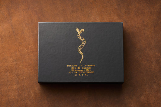 A black box adorned with a gold geometric snake design and gold lettering displaying “Persons of Interest Eau de Parfum Discovery Kit, 10 x 5ml Bottles” rests on a brown textured surface, inviting you to discover a curated collection of unisex fragrances from Persons of Interest.