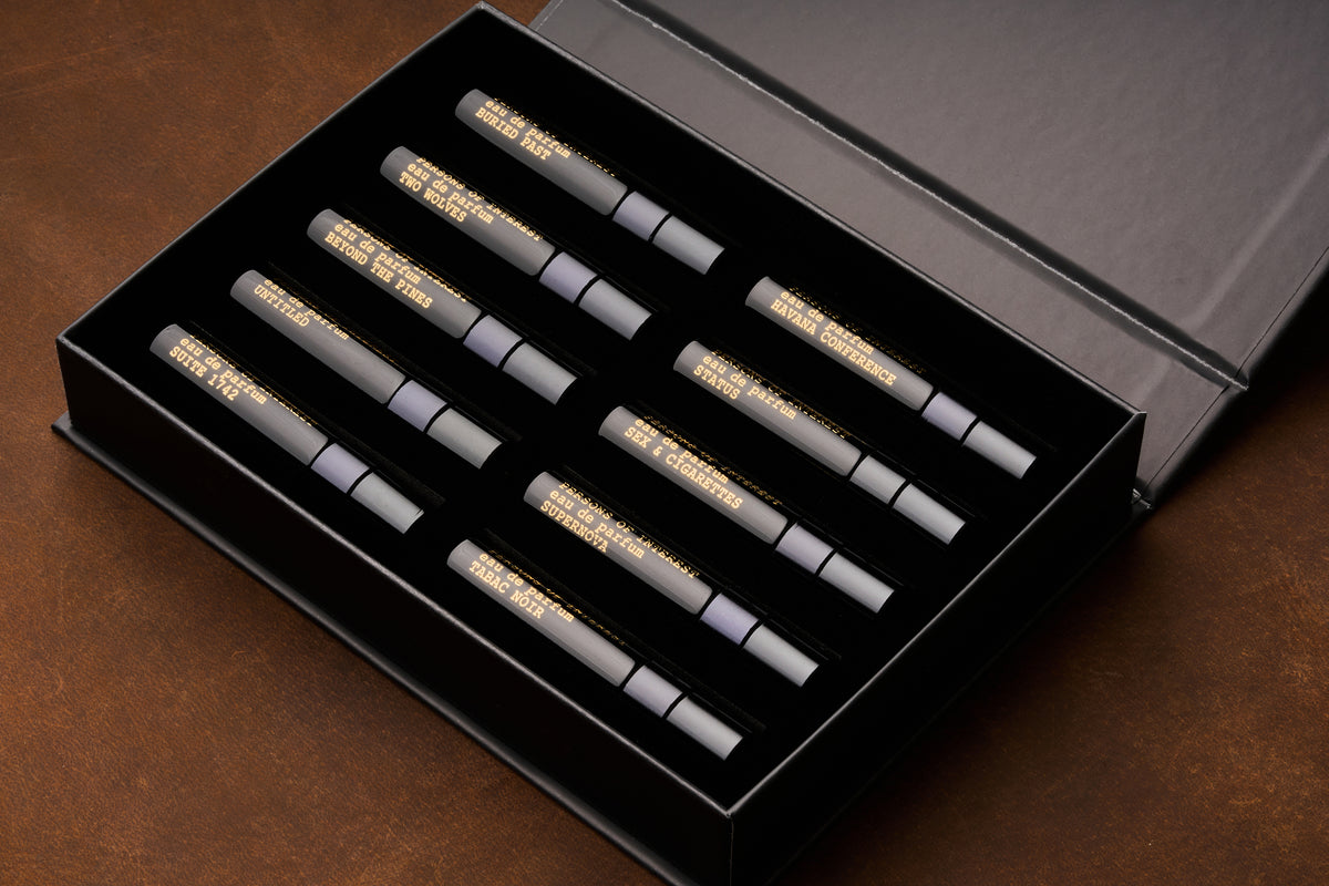 A black box containing ten 5ml perfume bottles with gold lettering, arranged in two rows on a brown surface—an elegant unisex fragrance discovery kit: the Persons of Interest Eau de Parfum Discovery Kit.