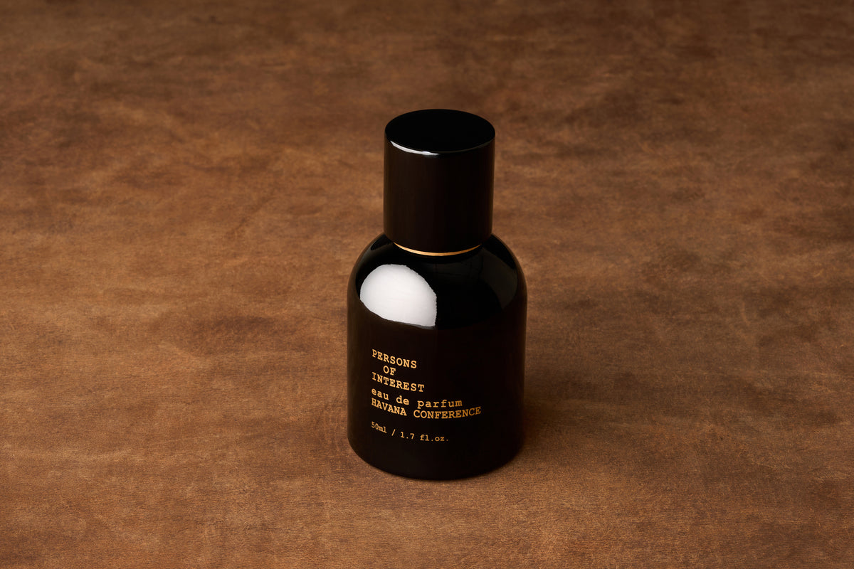 A black bottle of Persons of Interest Havana Conference Eau de Parfum rests on a brown textured surface, adorned with gold text on the front. This rum-scented fragrance exudes inviting warmth, making it ideal for those who appreciate a distinctive and memorable scent.