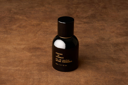 A black bottle of Persons of Interest Havana Conference Eau de Parfum rests on a brown textured surface, adorned with gold text on the front. This rum-scented fragrance exudes inviting warmth, making it ideal for those who appreciate a distinctive and memorable scent.
